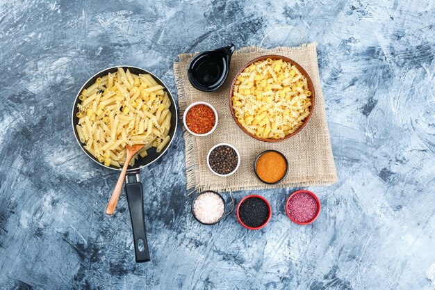 Conjunto de especias, cuchara, cuchara de madera y pasta cruda en una sartén y recipiente sobre yeso y un trozo de fondo de saco. vista superior.