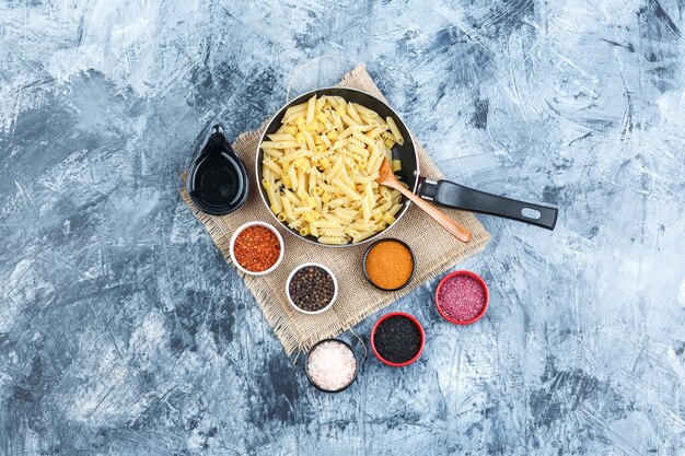 Conjunto de especias, cuchara, cuchara de madera y pasta cruda en una sartén sobre yeso y un trozo de fondo de saco. vista superior.