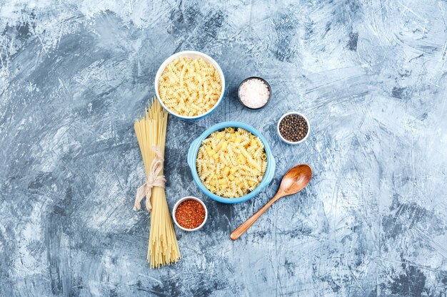 Conjunto de especias, cuchara de madera y pasta variada en tazones sobre un fondo de yeso gris. vista superior.