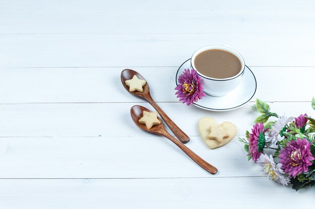 Conjunto de flores, galletas en cucharas de madera y taza de café, galletas en forma de corazón y estrella sobre un fondo de tablero de madera blanca. vista de ángulo alto.