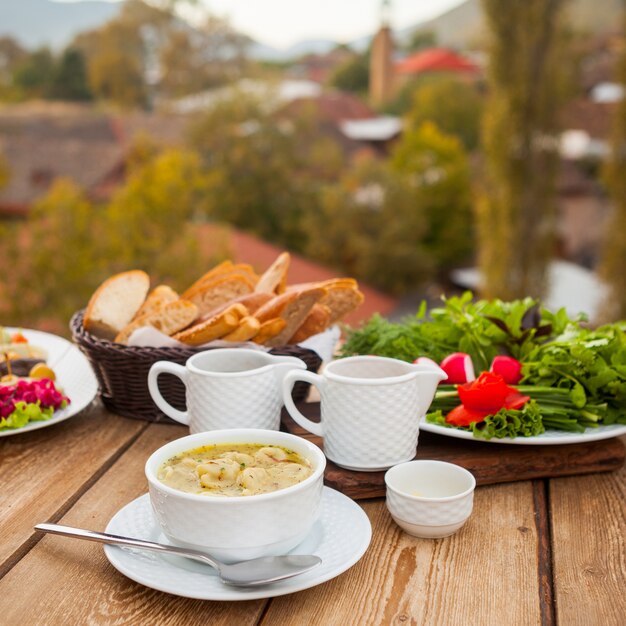 Conjunto de pan, verduras, ensalada y comida deliciosa sopa en un recipiente con una aldea en el fondo. Vista de ángulo alto.