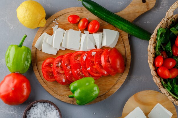 Conjunto de pimiento verde, limón, pepino, sal y queso en rodajas y tomates en una tabla de cortar sobre una superficie gris