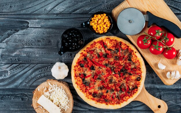 Conjunto de una rodaja de queso, ajo, tomate, aceitunas, maíz y un cortador de pizza y pizza en una tabla de pizza sobre un fondo de madera oscura. vista superior.