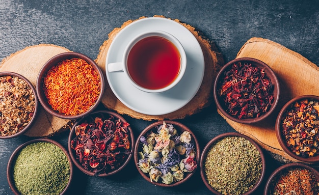 Conjunto de trozos de madera y una taza de té y hierbas de té en cuencos sobre un fondo oscuro con textura. aplanada