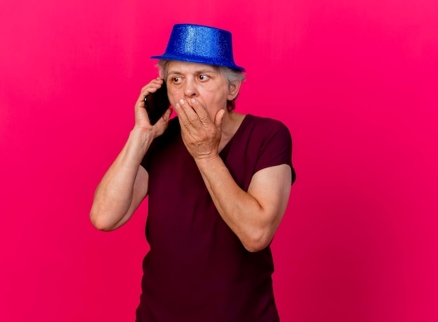 Conmocionada anciana con gorro de fiesta pone la mano en la boca hablando por teléfono en rosa