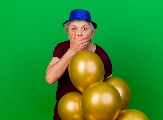 Conmocionada anciana con gorro de fiesta pone la mano en la boca de pie con globos de helio en verde