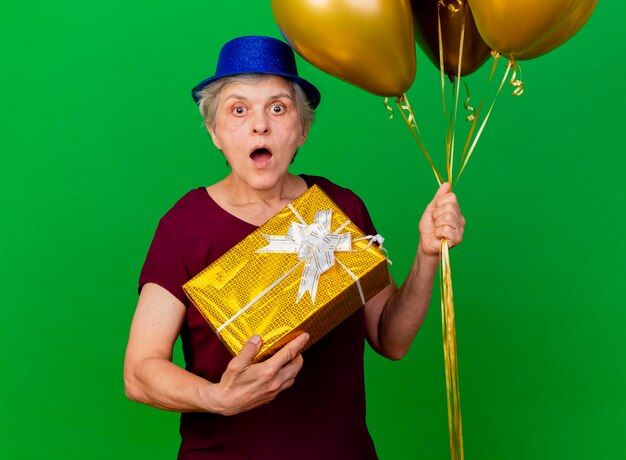 Conmocionada anciana con sombrero de fiesta sostiene globos de helio y caja de regalo en verde