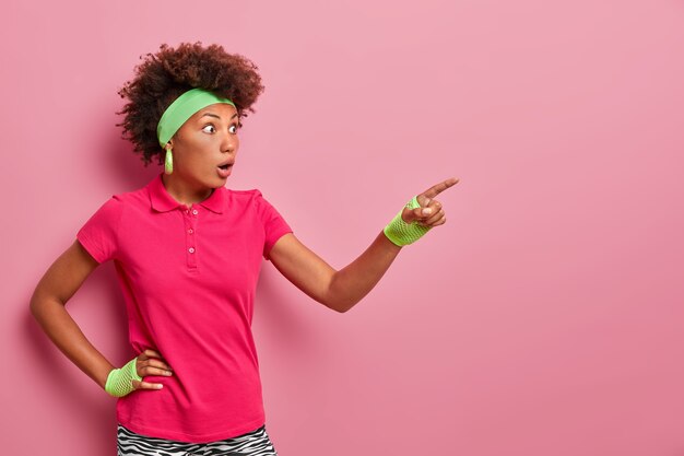 Conmocionada asombrada joven de pelo rizado tiene ojos saltones, señala con el dedo a la distancia, ve algo increíble, mira con la boca abierta, vestida con ropa deportiva