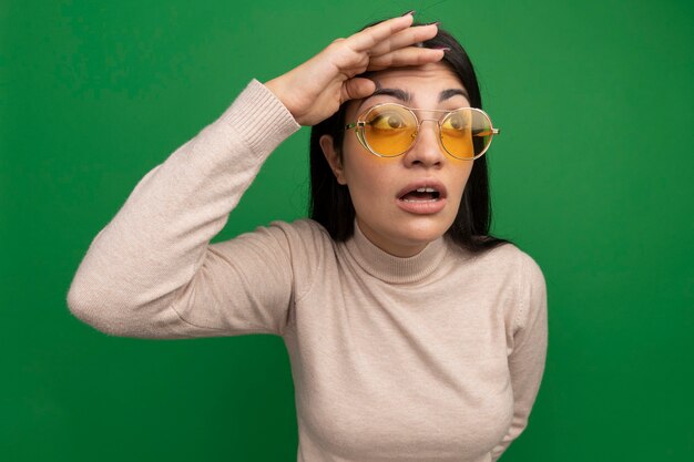 Conmocionada bastante morena chica caucásica en gafas de sol mantiene la palma en la frente mirando al lado en verde