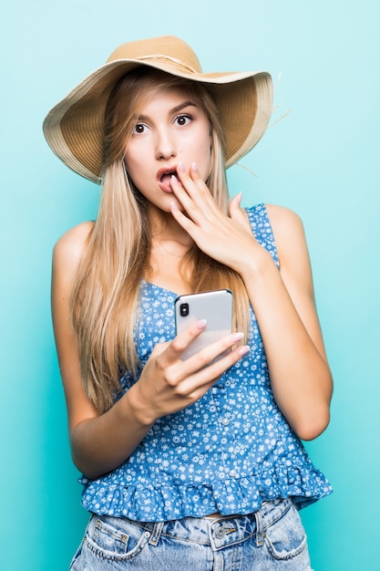 Conmocionada bastante morena mujer en vestido y sombrero de paja peleas por teléfono inteligente mientras mira a otro lado sobre fondo azul.