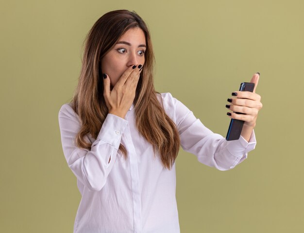 Conmocionada joven bastante caucásica pone la mano en la boca sosteniendo y mirando el teléfono