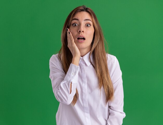 Conmocionada joven bastante caucásica pone la mano en la cara y mira a la cámara en verde