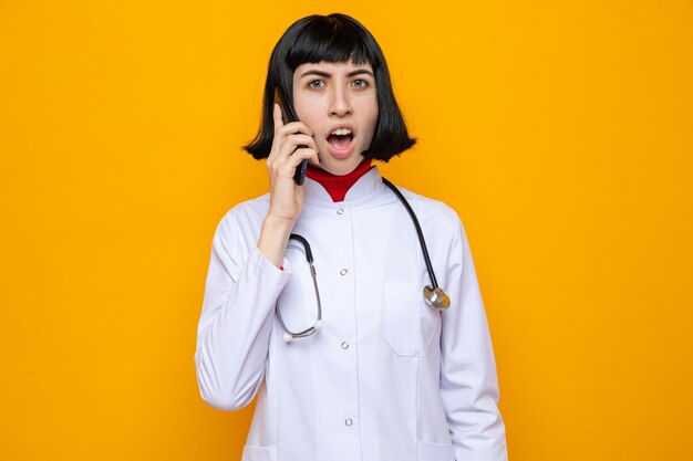 Conmocionada joven bastante caucásica en uniforme médico con estetoscopio hablando por teléfono