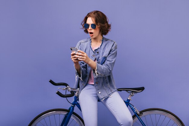 Conmocionada joven con cabello castaño mirando la pantalla del teléfono. Espectacular modelo de mujer sentada en bicicleta y usando su celular.