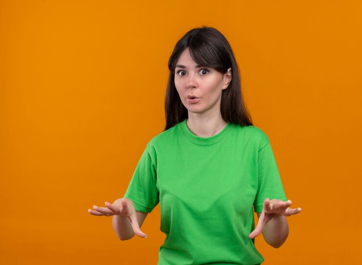 Foto gratuito conmocionada joven caucásica en camisa verde tiene las manos hacia abajo y mira a la cámara sobre fondo naranja aislado con espacio de copia