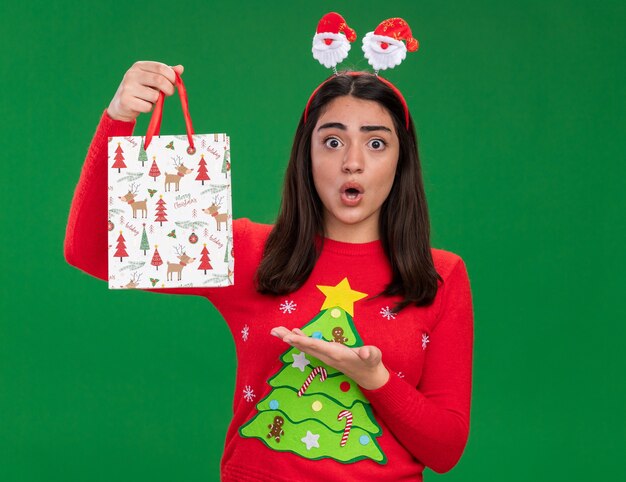 Conmocionada joven caucásica con diadema de santa sostiene y apunta a una bolsa de regalo de papel aislado sobre fondo verde con espacio de copia