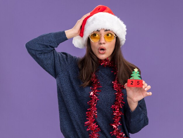 Conmocionada joven caucásica en gafas de sol con gorro de Papá Noel y guirnalda alrededor del cuello sostiene el adorno del árbol de Navidad y pone la mano en la cabeza mirando hacia el lado aislado sobre fondo púrpura con espacio de copia