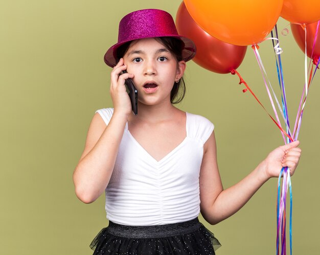 Conmocionada joven caucásica con gorro de fiesta púrpura sosteniendo globos de helio hablando por teléfono aislado en la pared verde oliva con espacio de copia