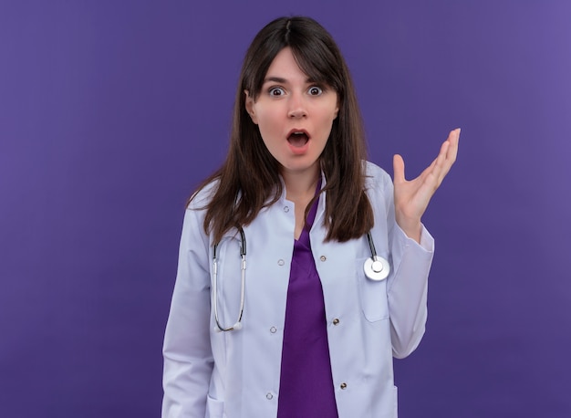 Conmocionada joven doctora en bata médica con estetoscopio levanta la mano sobre fondo violeta aislado con espacio de copia