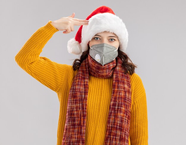 Conmocionada joven eslava con gorro de Papá Noel y con bufanda alrededor del cuello con máscara médica haciendo gesto de pistola y poniendo en su templo aislado sobre fondo blanco con espacio de copia