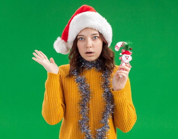 Conmocionada joven eslava con gorro de Papá Noel y con guirnalda alrededor del cuello sosteniendo bastón de caramelo y manteniendo la mano abierta