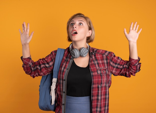 Conmocionada joven estudiante eslava con auriculares con mochila se encuentra con las manos levantadas