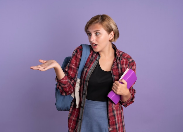 Conmocionada joven estudiante eslava vestida con mochila sostiene el libro y el cuaderno mirando y apuntando al lado