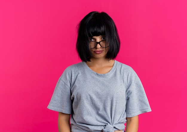 Conmocionada joven morena caucásica en gafas ópticas mira a cámara aislada sobre fondo rosa con espacio de copia