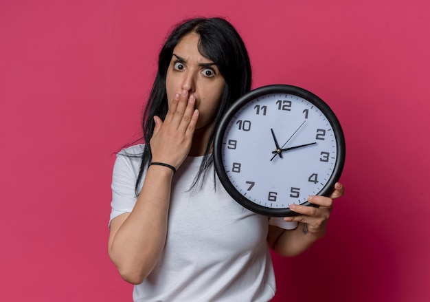 Foto gratuita conmocionada joven morena caucásica pone la mano en la boca y sostiene el reloj aislado en la pared rosa