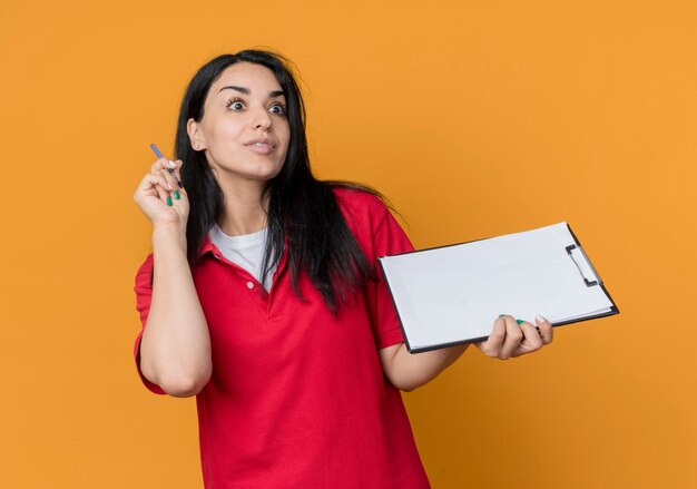 Conmocionada joven morena caucásica vestida con camisa roja sostiene la pluma y el portapapeles mirando hacia adelante aislado en la pared naranja