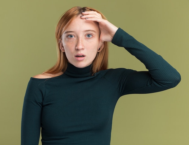 Conmocionada joven pelirroja pelirroja con pecas pone la mano en la cabeza en verde oliva