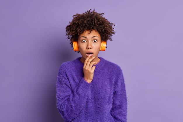 Conmocionada joven de pelo rizado con cabello afro mira sorprendentemente a la cámara, mantiene la boca abierta, usa auriculares estéreo, suéter púrpura, escucha noticias impactantes en poses de radio en interiores Concepto de Omg