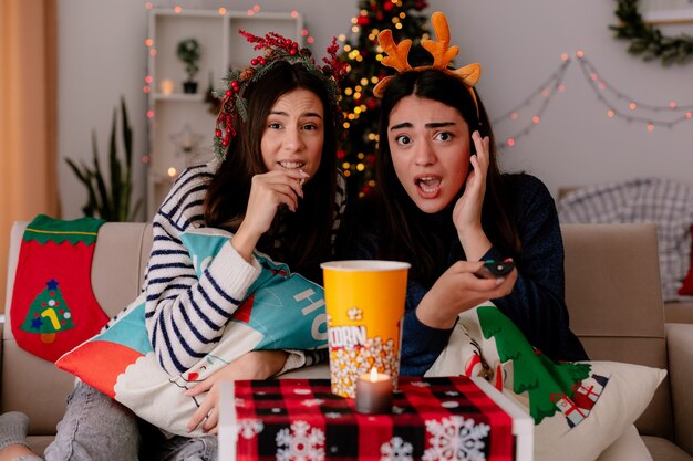 Conmocionada jovencita con diadema de renos sostiene el control remoto de la televisión sentada en un sillón con su amiga comiendo palomitas de maíz disfrutando de la Navidad en casa