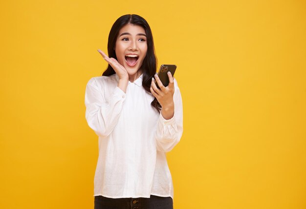 Conmocionada mujer asiática con camiseta blanca informal hablando por teléfono móvil en un estudio de fondo amarillo