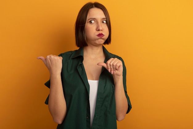 Conmocionada mujer bastante caucásica sopla mejillas mirando y apuntando al lado con las dos manos en naranja