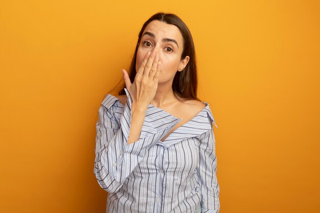 Conmocionada mujer bonita pone la mano en la boca aislada en la pared naranja