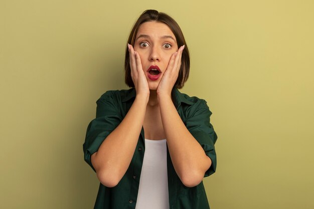 Conmocionada mujer bonita pone las manos en la cara aislada en la pared verde oliva