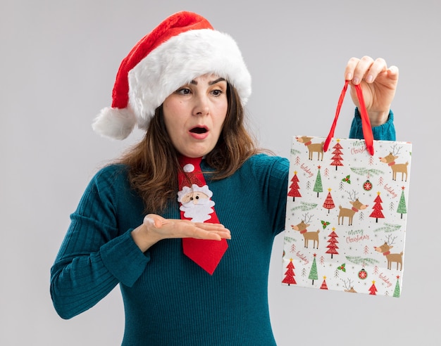 Conmocionada mujer caucásica adulta con gorro de Papá Noel y corbata de santa mirando y apuntando a la caja de regalo de papel aislada en la pared blanca con espacio de copia