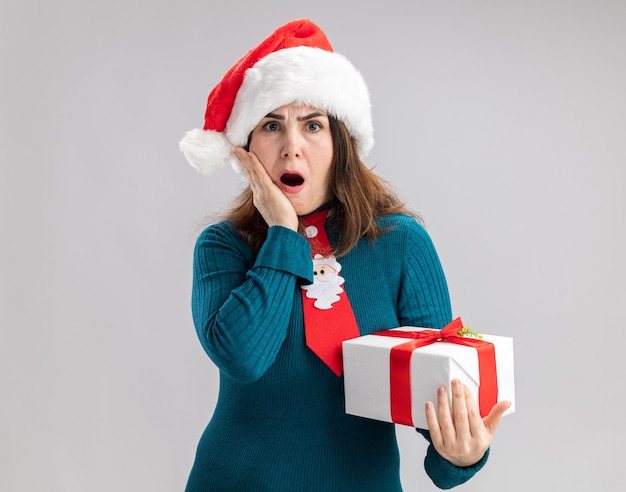 Conmocionada mujer caucásica adulta con gorro de Papá Noel y corbata de santa pone la mano en la cara y sostiene la caja de regalo de Navidad aislada en la pared blanca con espacio de copia