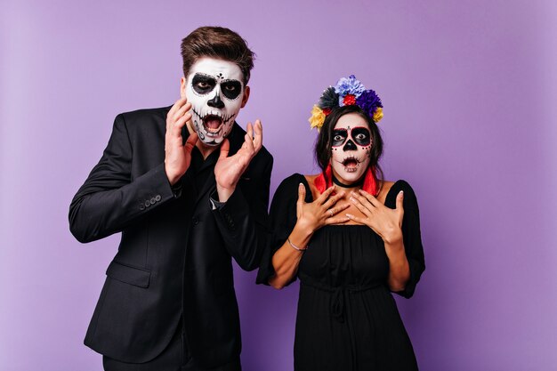 Conmocionada mujer caucásica con cabello negro posando en traje de halloween. Chico zombie sorprendido de pie en la pared púrpura con su novia.