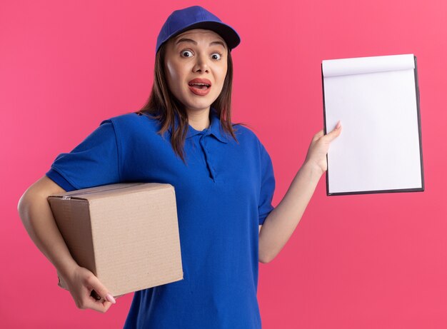 Conmocionada mujer de entrega bonita en uniforme sostiene cardbox y portapapeles aislado en la pared rosa con espacio de copia