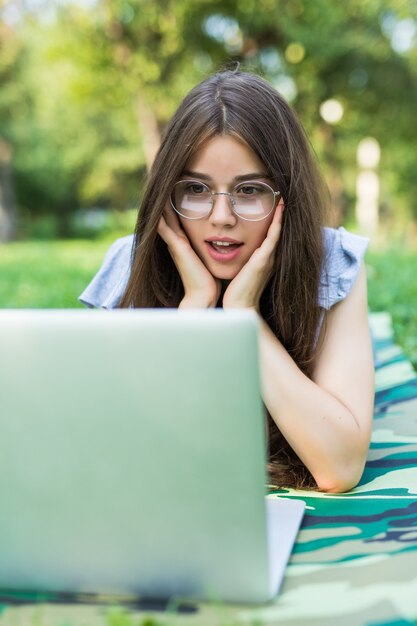 Conmocionada mujer morena en anteojos tumbado en la hierba en el parque y usando la computadora portátil