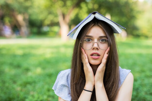 Conmocionada mujer morena sentada en el parque con el libro en la cabeza mientras sostiene sus brazos en las mejillas y mirando a la cámara