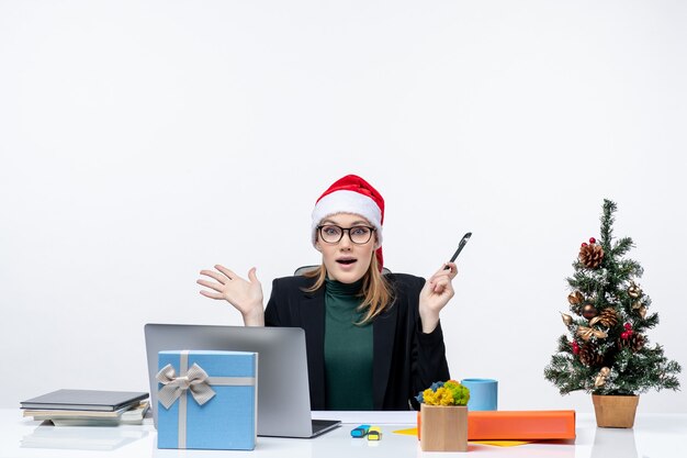 Conmocionada mujer rubia con un sombrero de santa claus sentado en una mesa con un árbol de Navidad y un regalo en la oficina sobre fondo blanco.