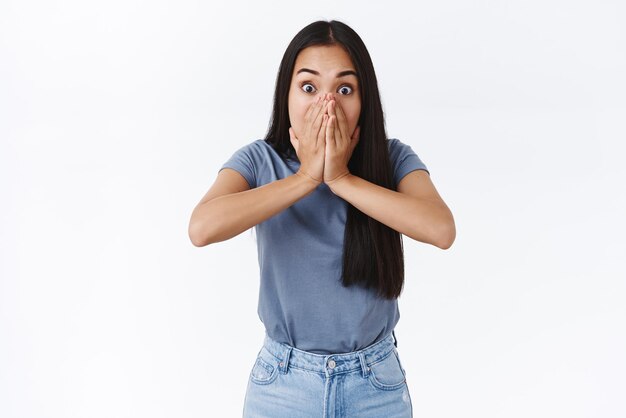 Conmocionada, sin palabras, una chica morena asiática mirando a la cámara, asombrada, jadeando, sostenga su boca, no grite por la sacudida y el asombro, sea testigo de algo impresionante, soporte, fondo blanco, concepto de emociones.