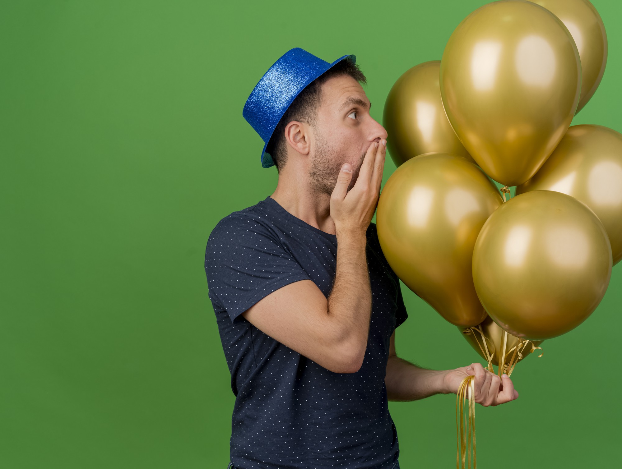 Conmocionado apuesto hombre caucásico vestido con gorro de fiesta azul sostiene globos de helio pone la mano en la boca aislada sobre fondo verde con espacio de copia