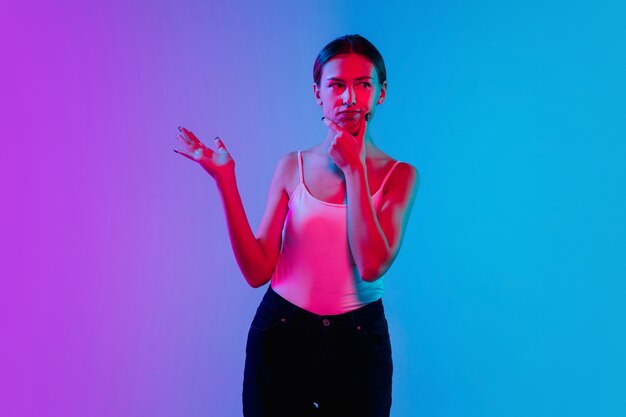 Conmocionado, asombrado. Retrato de joven mujer caucásica sobre fondo de estudio azul-púrpura degradado en luz de neón. Concepto de juventud, emociones humanas, expresión facial, ventas, publicidad. Preciosa modelo morena.