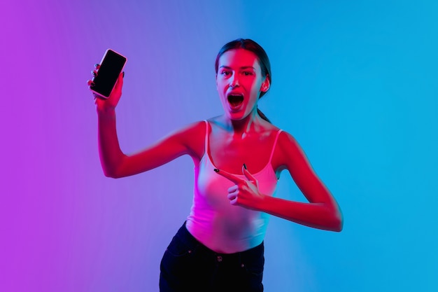 Conmocionado, asombrado. Retrato de joven mujer caucásica sobre fondo de estudio azul-púrpura degradado en luz de neón. Concepto de juventud, emociones humanas, expresión facial, ventas, publicidad. Preciosa modelo morena.