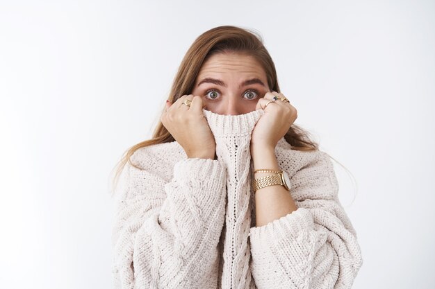 Conmocionado asustado aturdido linda mujer caucásica escondiendo suéter de cuello de cara tirando de tela nariz ensanchar los ojos asombrado sin palabras asustado viendo horror de pie aturdido fondo blanco, asustado