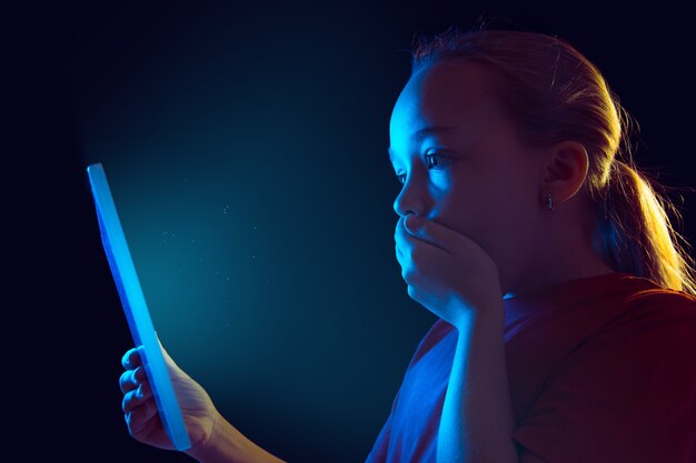 Conmocionado, asustado. Retrato de niña caucásica sobre fondo oscuro de estudio en luz de neón. Modelo femenino hermoso que usa la tableta. Concepto de emociones humanas, expresión facial, ventas, publicidad, tecnología moderna, gadgets.